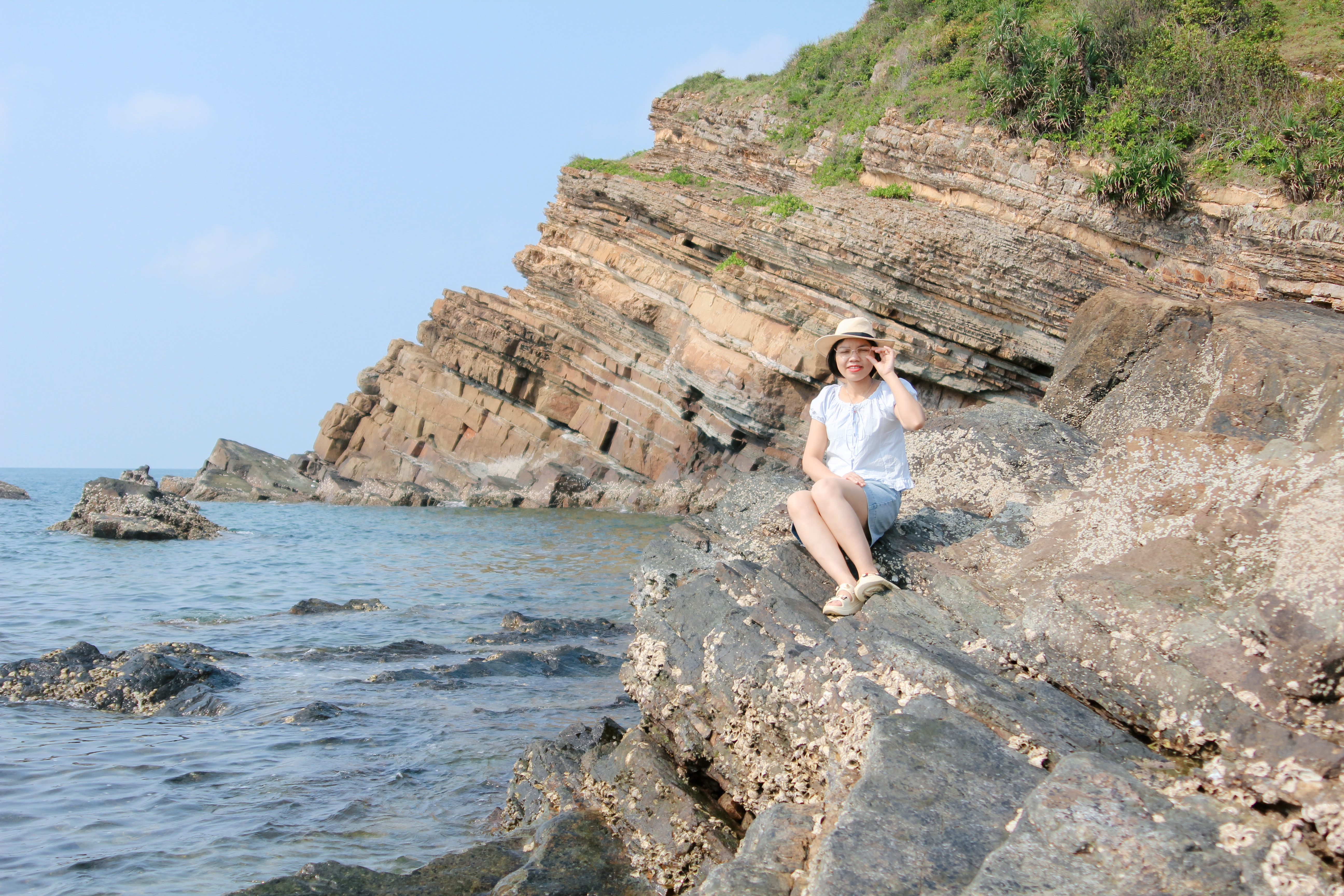A woman in a sunhat sits on a rocky shore enjoying a sunny day by the sea.