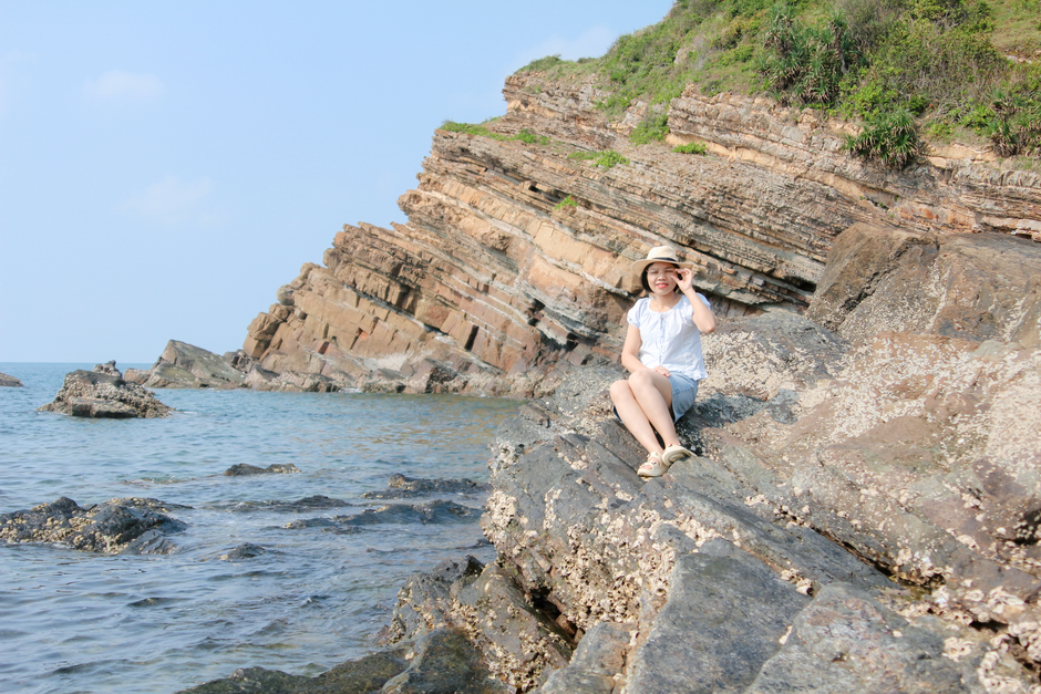 A woman in a sunhat sits on a rocky shore enjoying a sunny day by the sea.