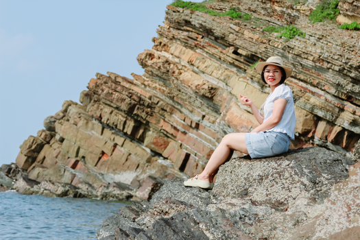 Woman sitting on a scenic rocky cliff overlooking the ocean on a sunny day, wearing casual summer attire.