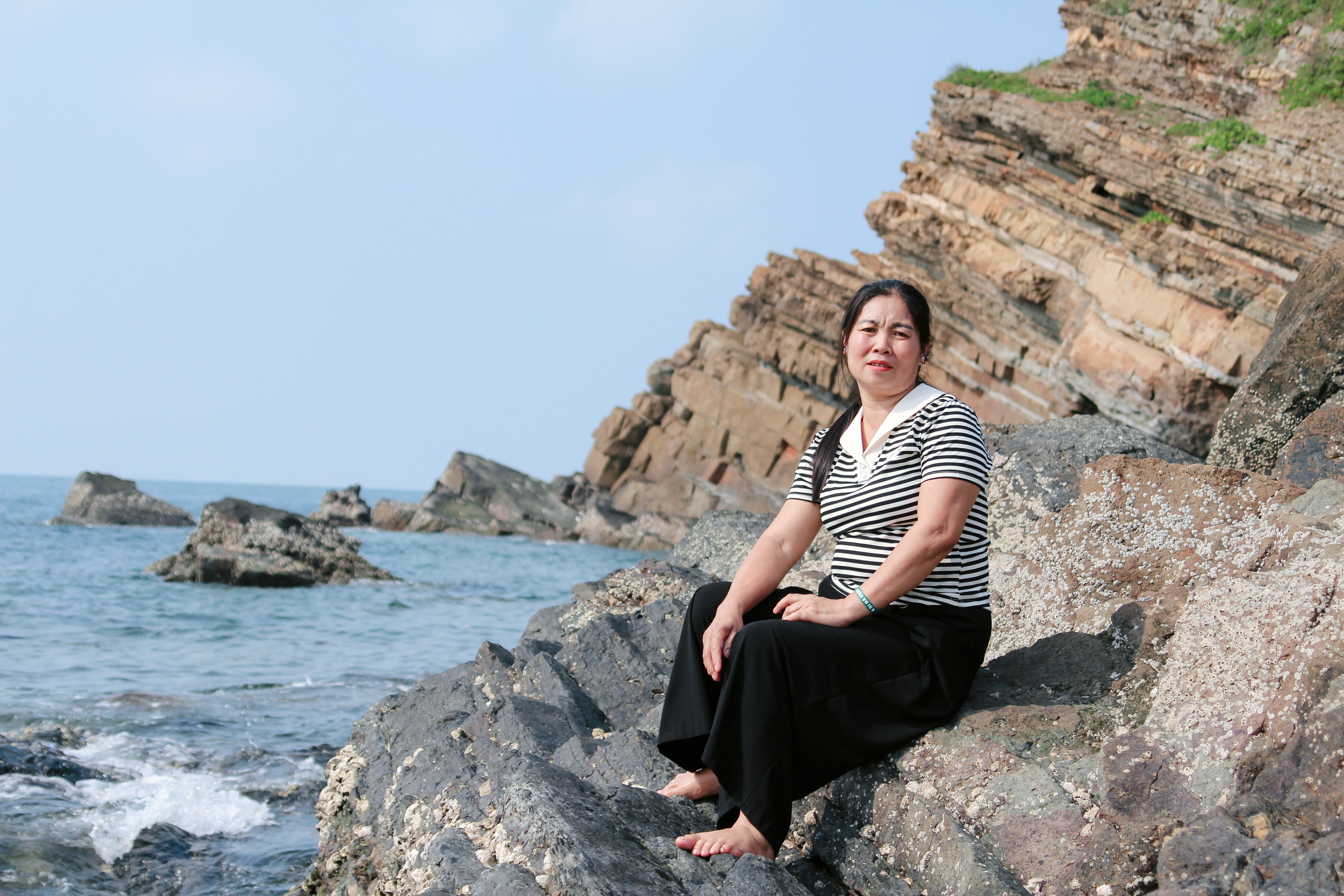 A woman sits on rocky cliffs by the sea, enjoying a peaceful day outdoors.