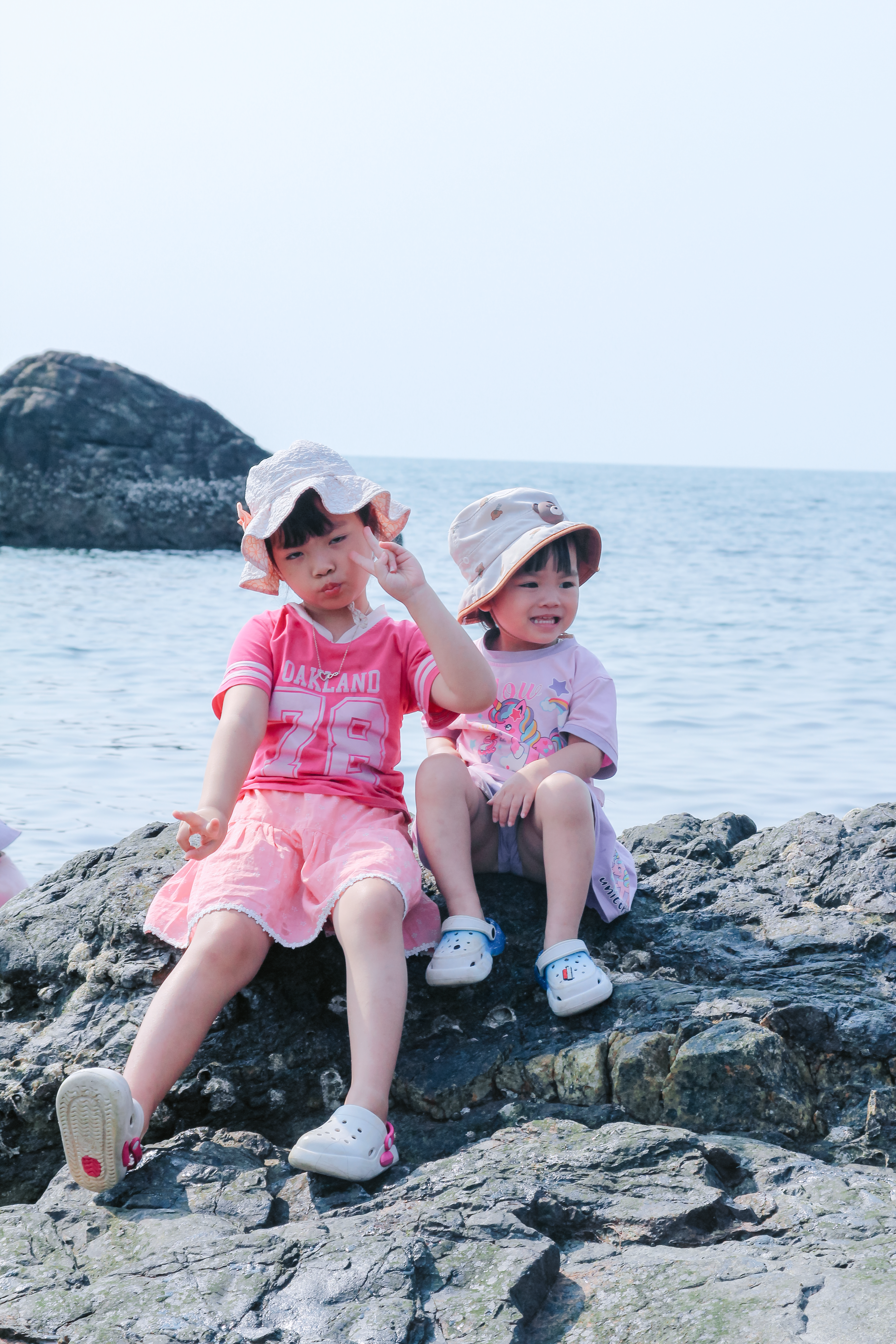 Two young children in hats enjoy a sunny day on rocky ocean shore.