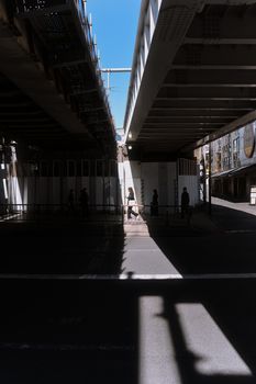 Silhouetted pedestrians walk under a Tokyo overpass, casting dramatic shadows in the sunlight.