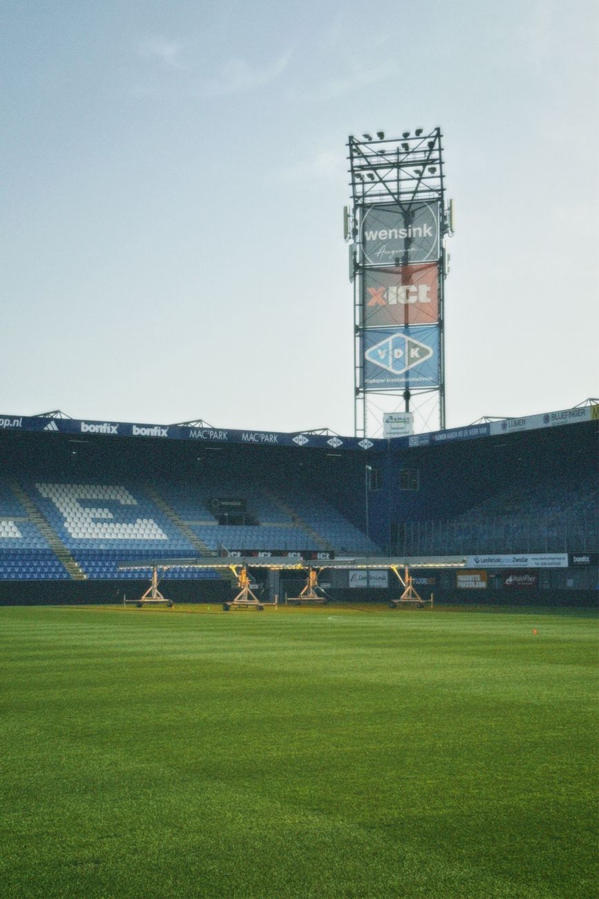 Een stil en leeg voetbalstadion met een heldergroen veld en torenhoge schijnwerpers onder een strakke hemel.