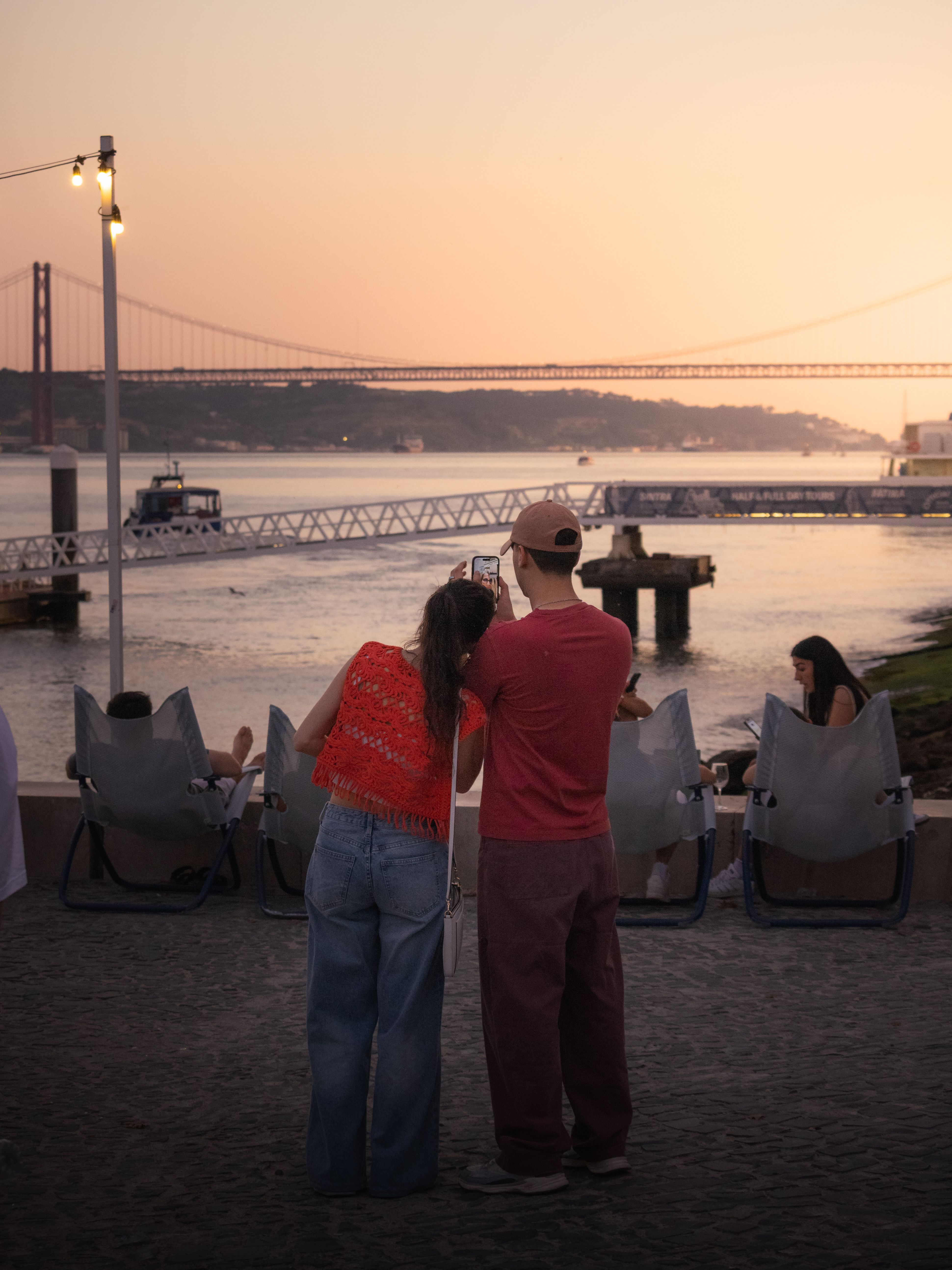A couple taking a photo with a scenic view of the Tagus River and bridge in Lisbon at sunset.