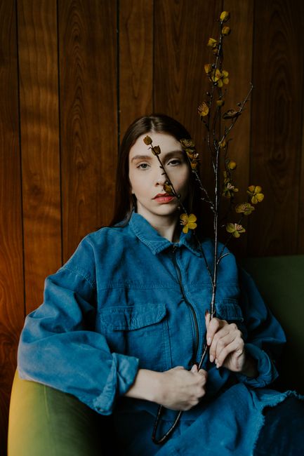 Artistic portrait of a woman in denim with a floral branch against a wooden backdrop, embodying a retro and vogue aesthetic.