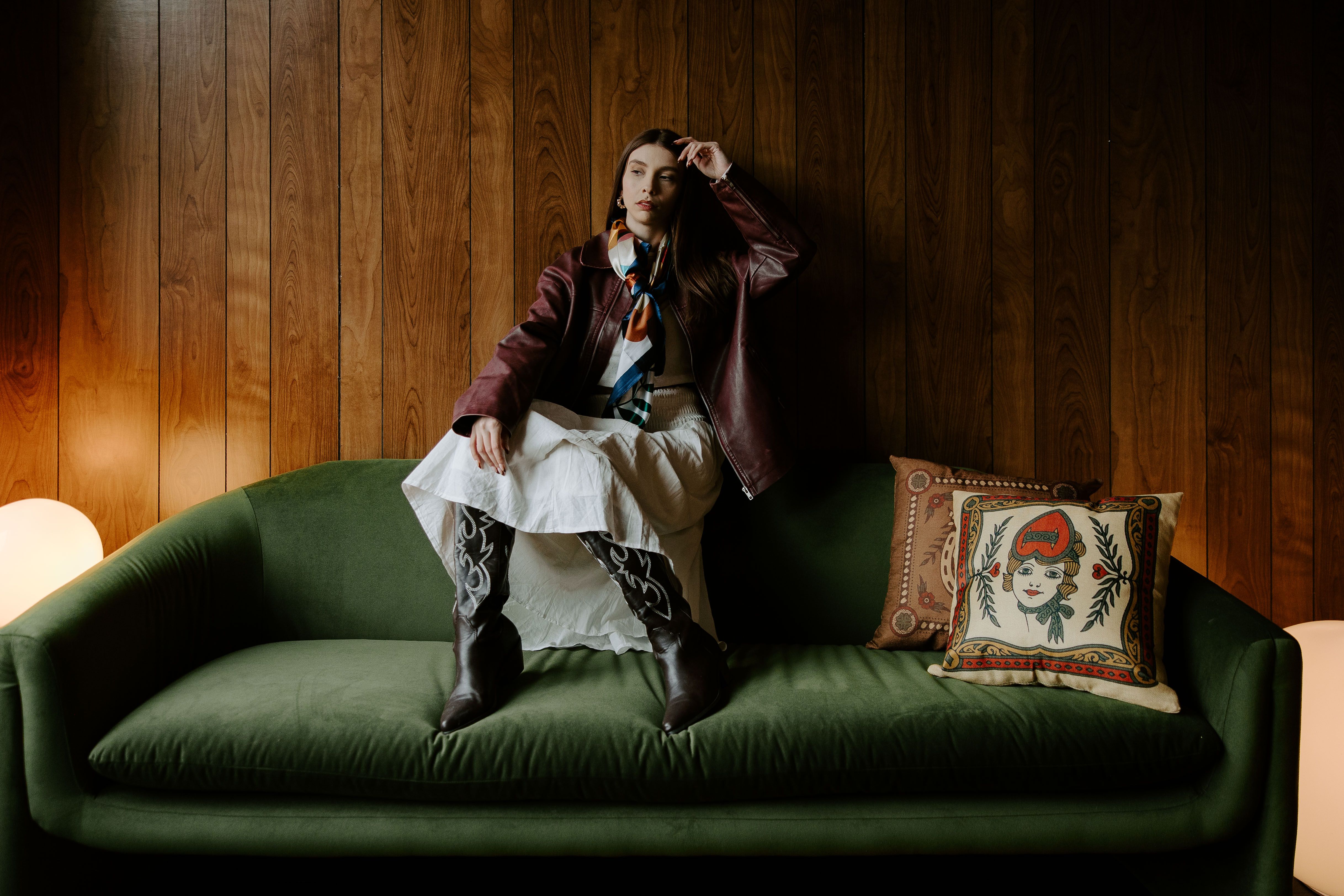 A fashionable woman in a leather jacket and boots sitting on a green sofa indoors, exuding elegance and style.