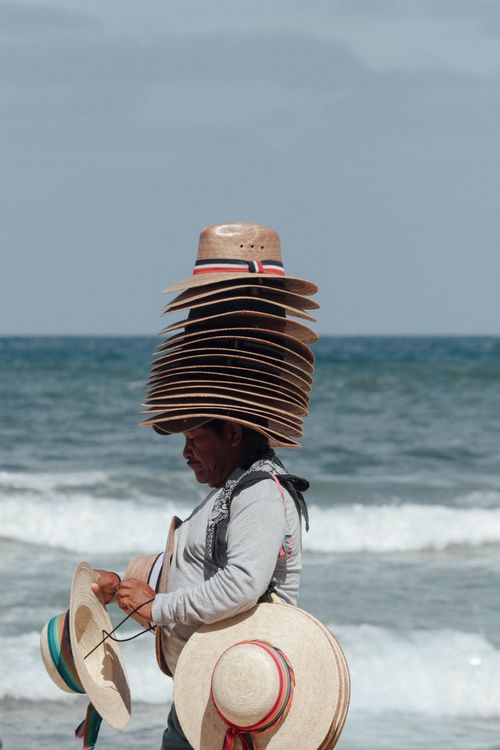 Free A street vendor balancing multiple stacked straw hats on the beach, offering them for sale. Stock Photo