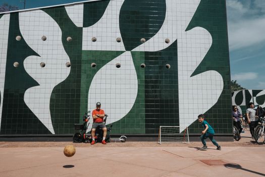 Child playing soccer in front of a modern artistic wall with an adult observing in a vibrant urban setting.
