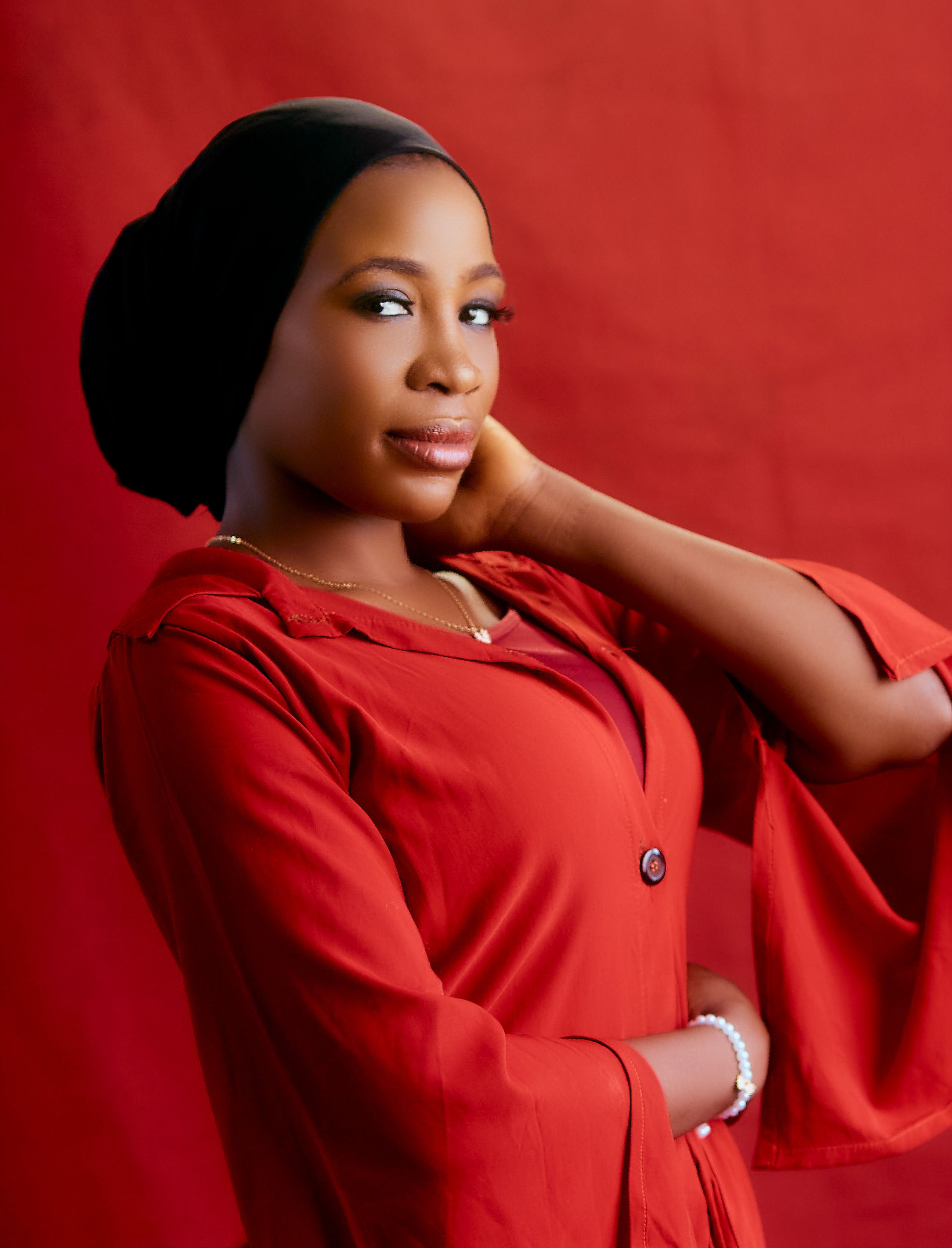 Elegant woman in red clothing with black headscarf posing against a red backdrop.