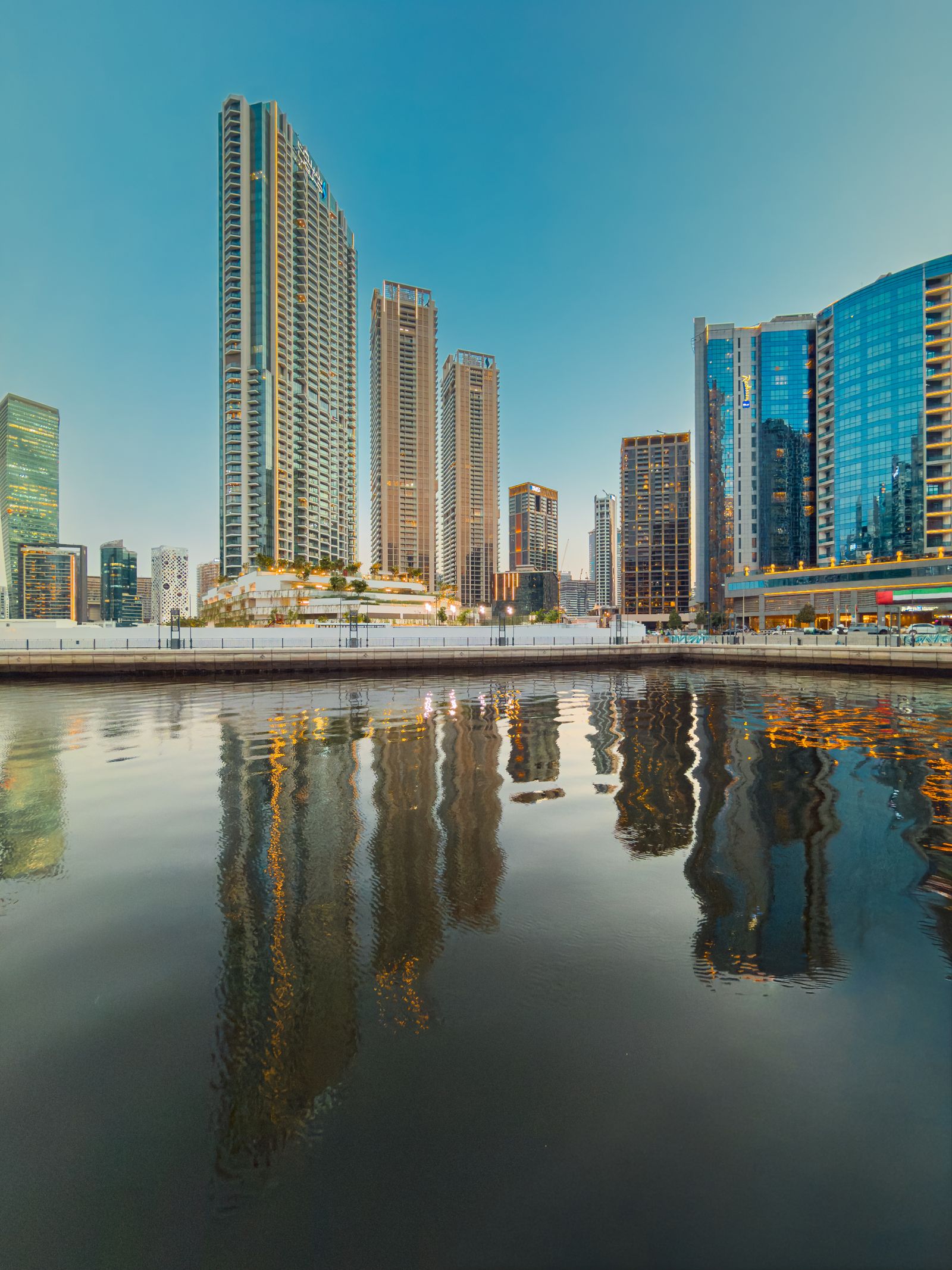 Business Bay towers along the Dubai Water Canal reflected in the still water at dusk, including the canal-side waterfront hotel.