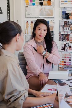 Two women discuss fashion designs in an office with mood boards and fabric samples.