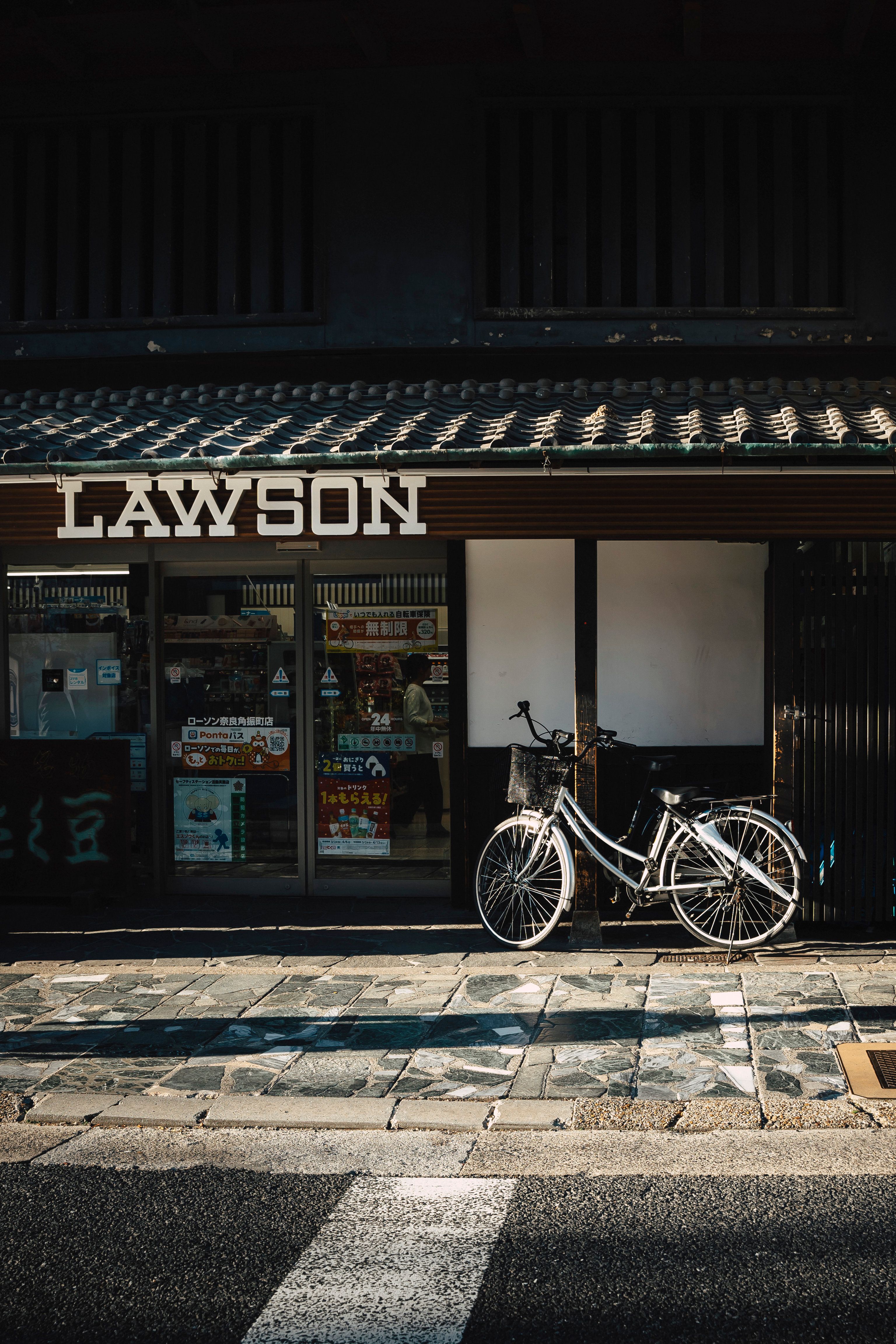 Bicycle parked outside a Lawson convenience store with a traditional facade, shadowed street view.