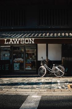 Bicycle parked outside a Lawson convenience store with a traditional facade, shadowed street view.