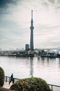 View of Tokyo Skytree from a riverside with a person jogging by. Cloudy sky.