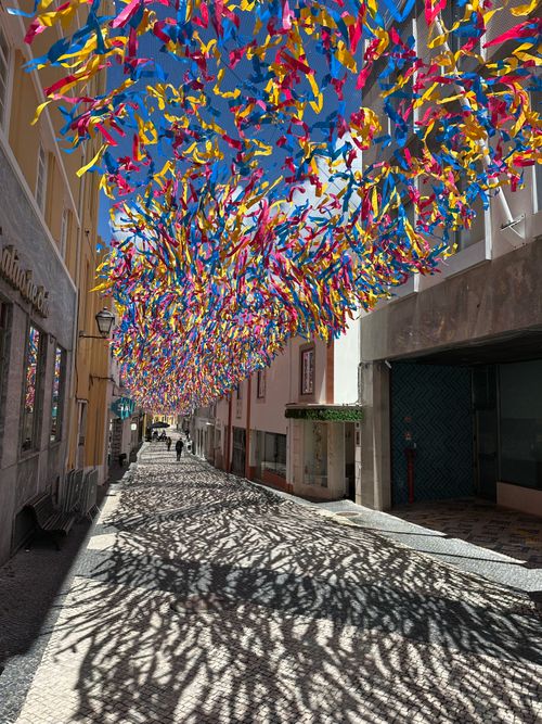 Free Colorful ribbons create a festive canopy over a sunny street in Torres Vedras, Portugal, casting vivid shadows. Stock Photo