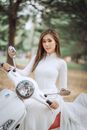 一位身着白色连衣裙的优雅女子，在风景优美的公园里骑着滑板车