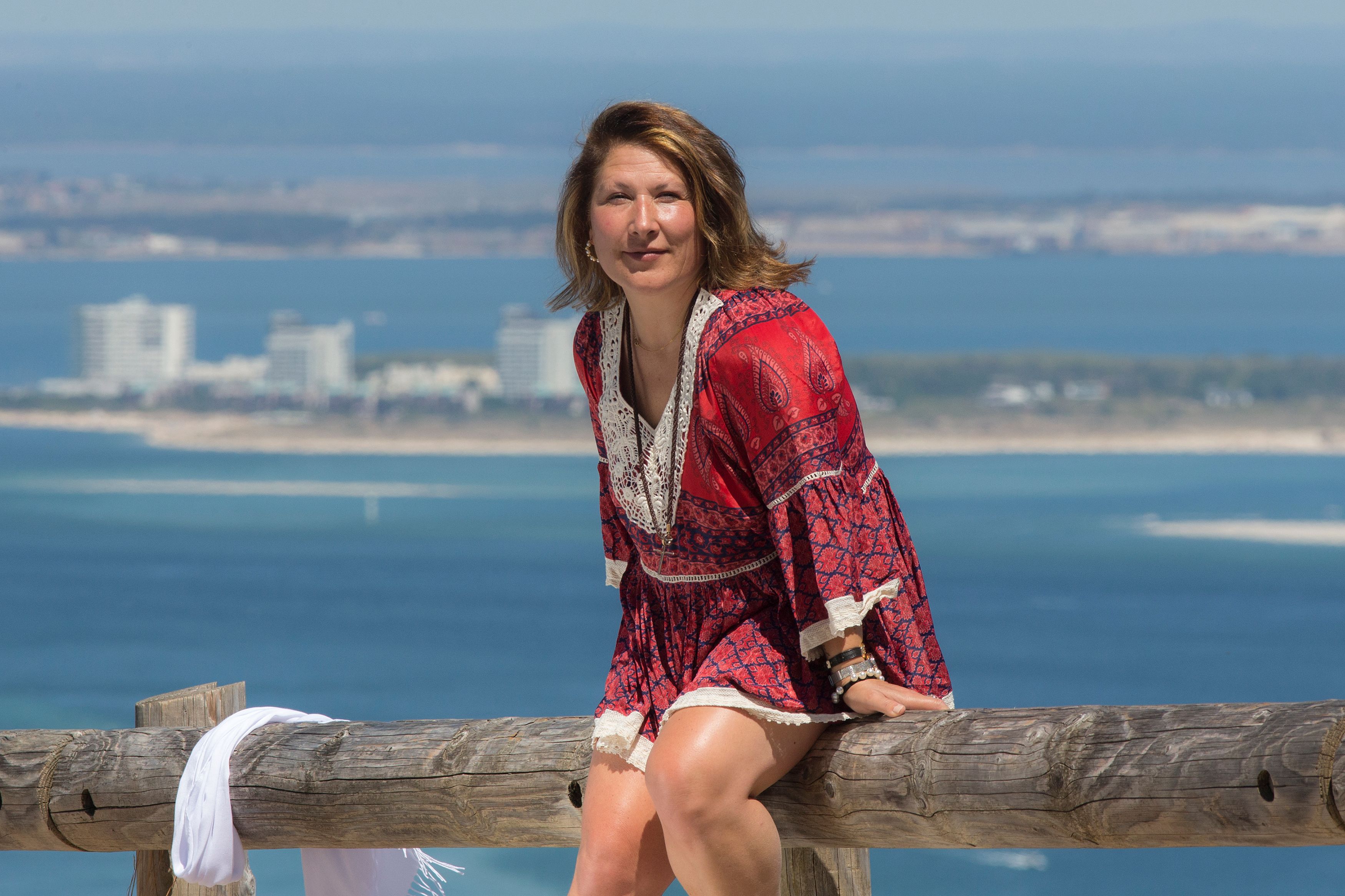 Woman in a red dress sitting on a fence with a coastal city view in the background.