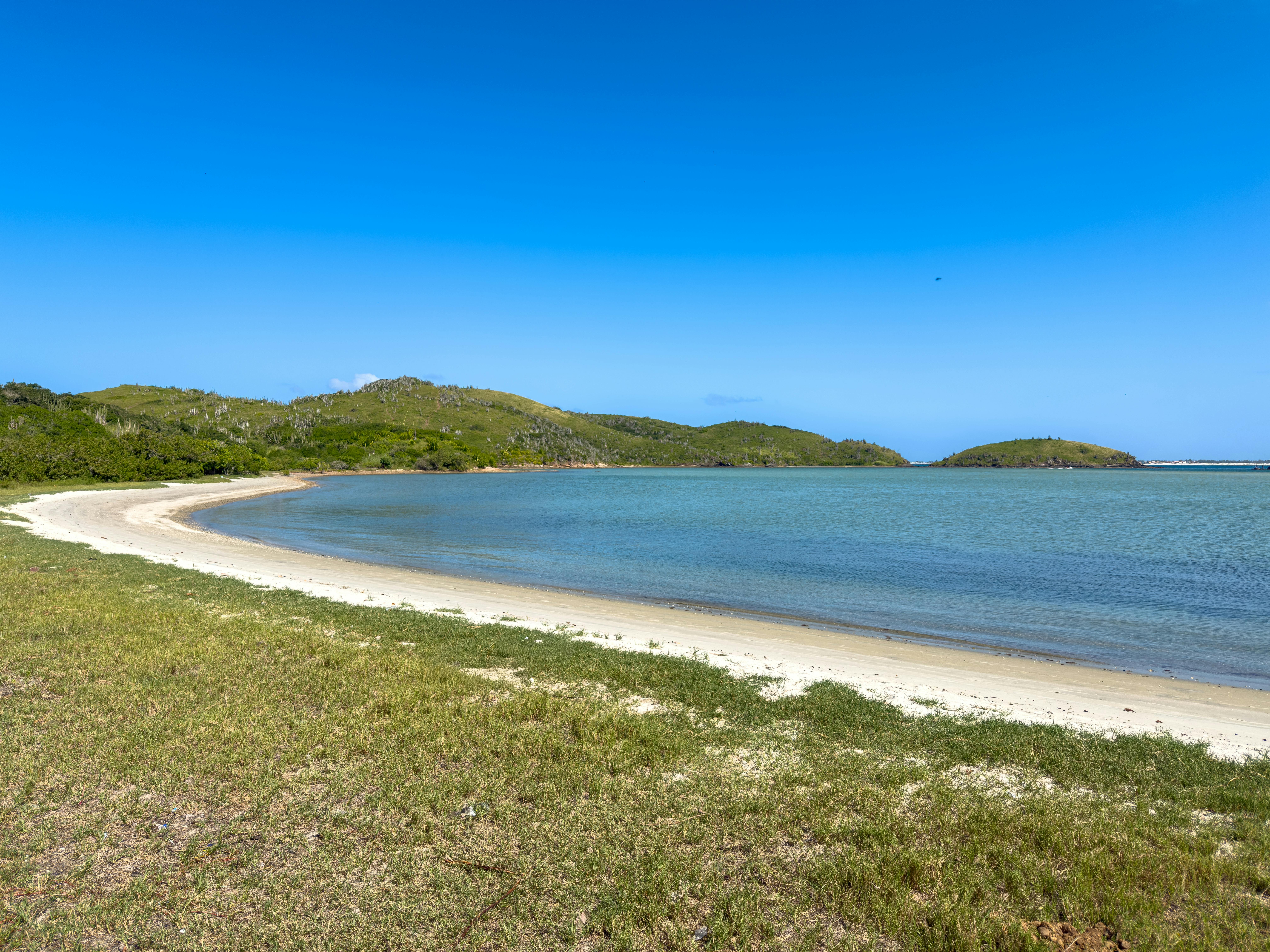 Idyllic beach scene with clear blue sky, turquoise water, and lush green hills in the background.