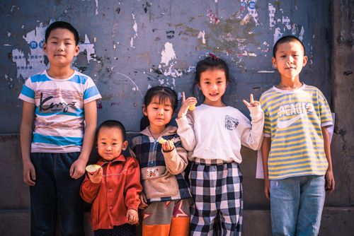 Free Five Asian children pose against a wall, showing playfulness and camaraderie in an outdoor setting. Stock Photo