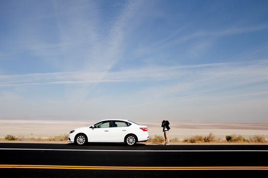 A white car parked on a desert road with a photographer capturing the vast landscape under a clear sky.