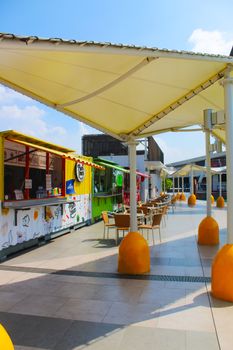 Colorful food stalls and seating area under canopied structure in a bright outdoor market setting.
