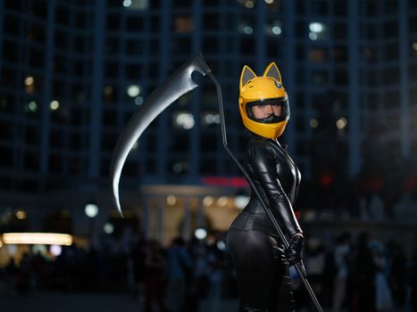 Cosplayer in Vietnam wearing black suit with yellow helmet holding scythe. Night city backdrop.