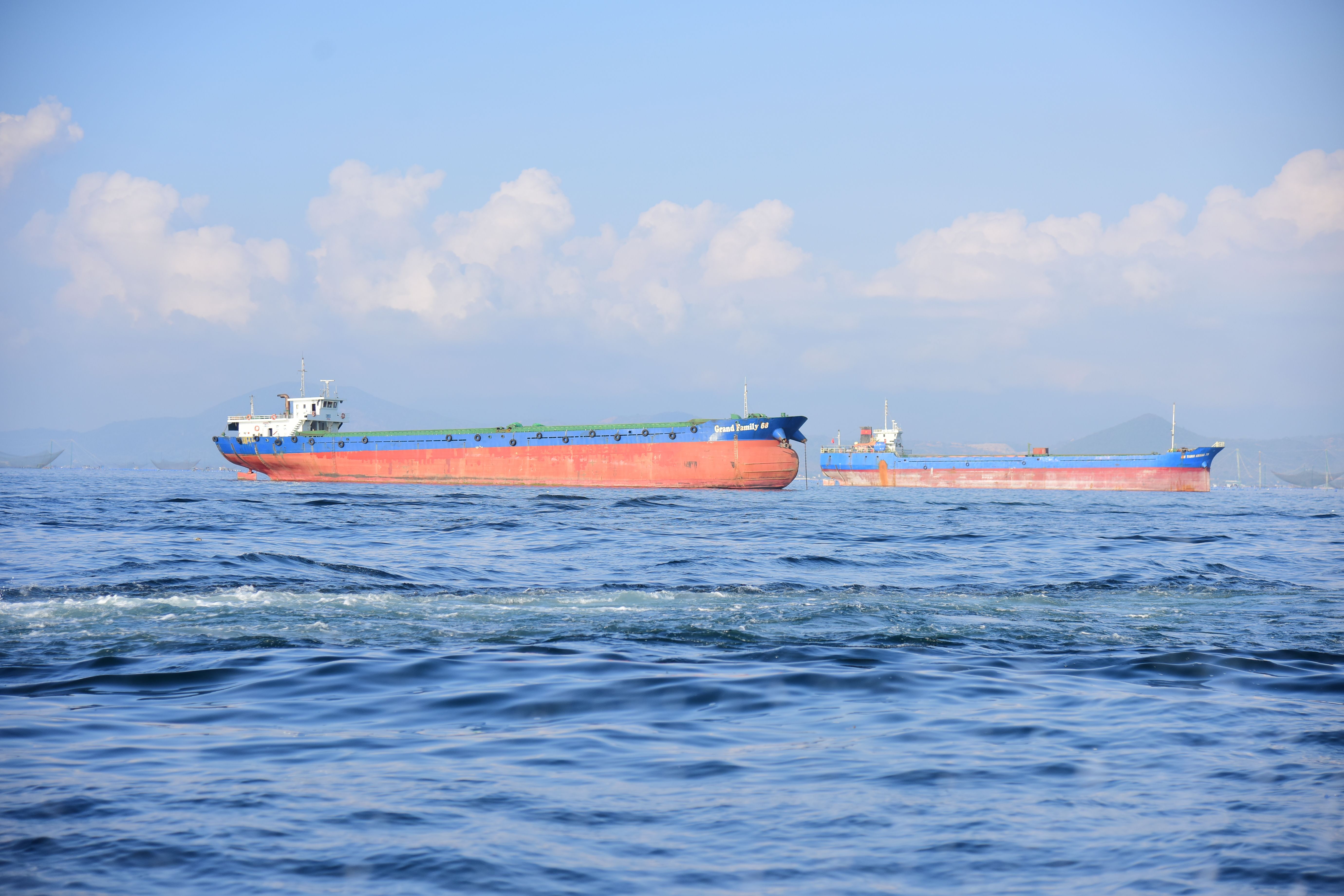Two large cargo ships sail on the ocean with the horizon and clouds in the background.