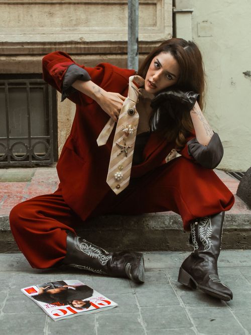 Free Fashion-forward woman in a bold red suit and boots, enhancing style with an embellished tie, sitting on a city street. Stock Photo