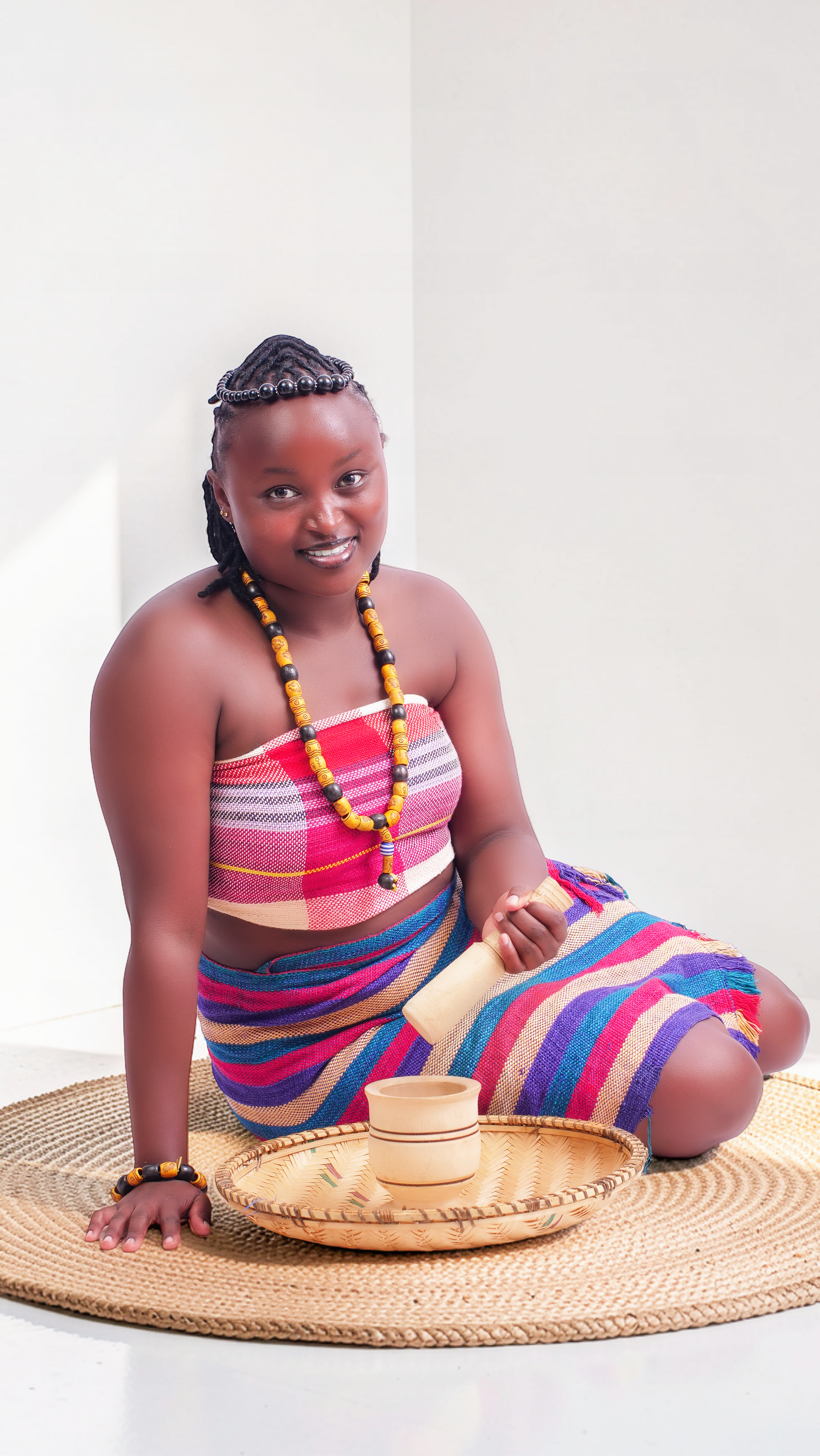 A smiling African woman in vibrant traditional dress seated indoors on a woven mat.