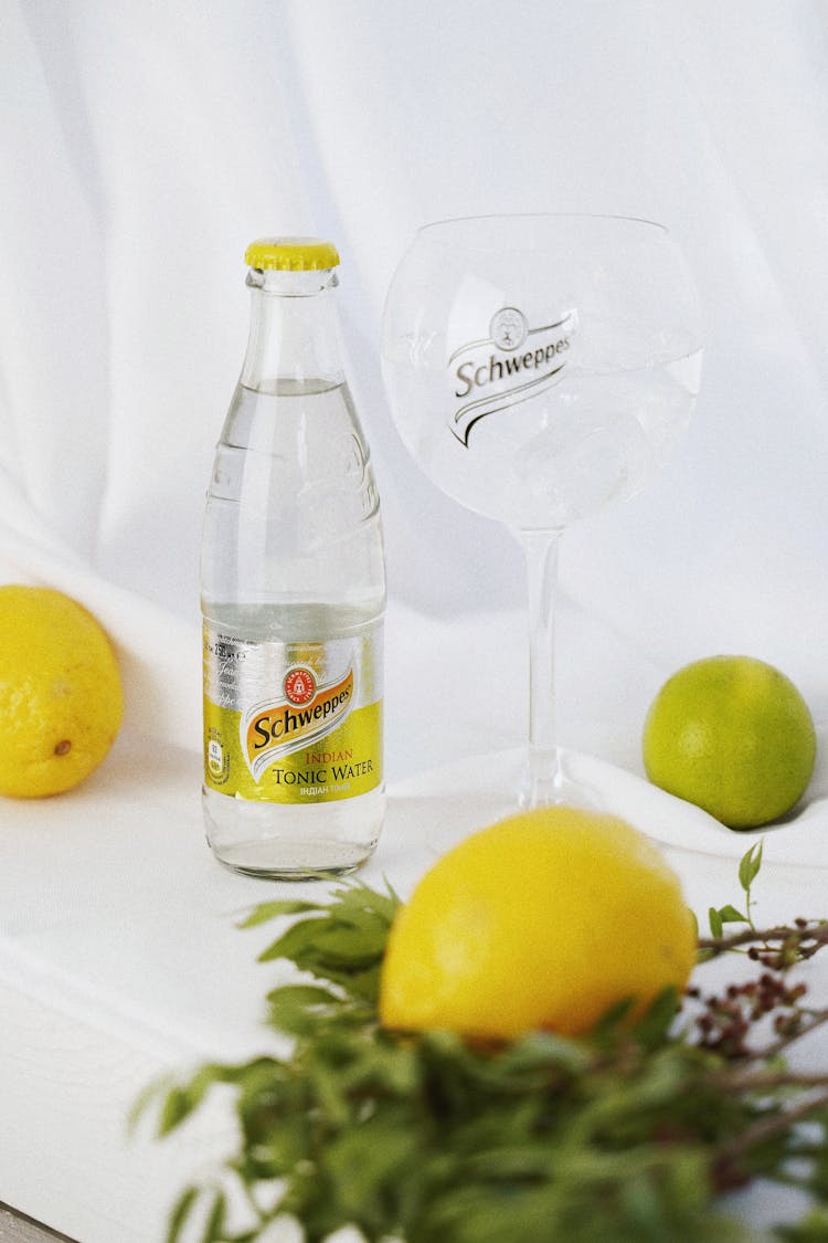 A Bottle Of Tonic Water Surrounded With Citrus Fruits On A White Surface