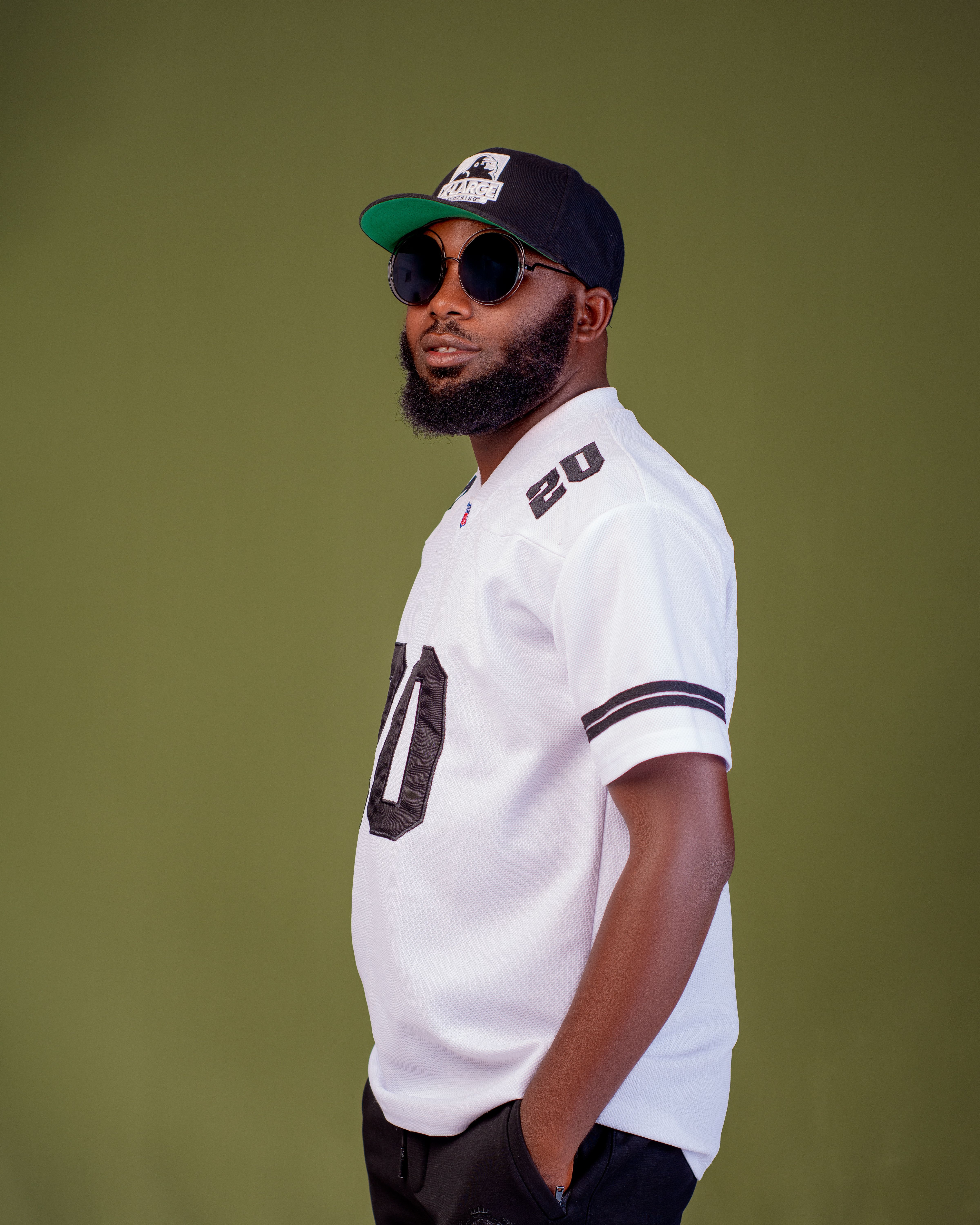 Man in sunglasses, baseball jersey, and cap against olive background.