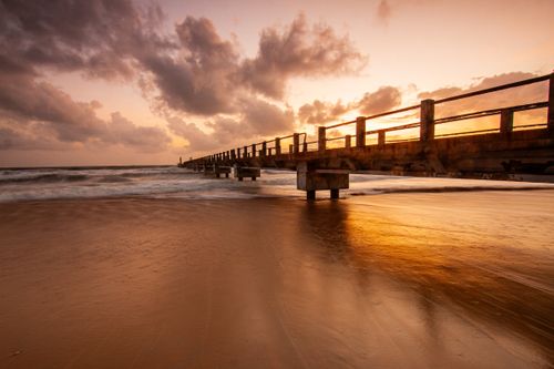 Free A stunning photograph of a beach pier at sunset with dramatic clouds and golden reflections on the water. Stock Photo