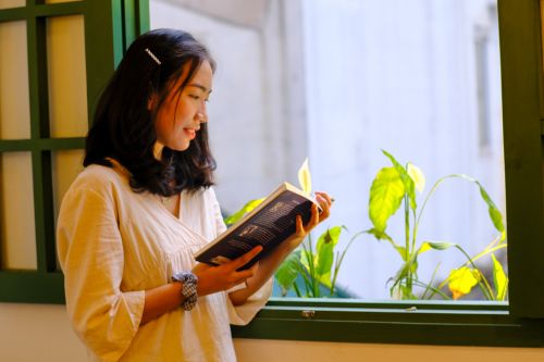 Free A young woman enjoys reading by a sunlit window, capturing a serene moment in Hue, Vietnam. Stock Photo