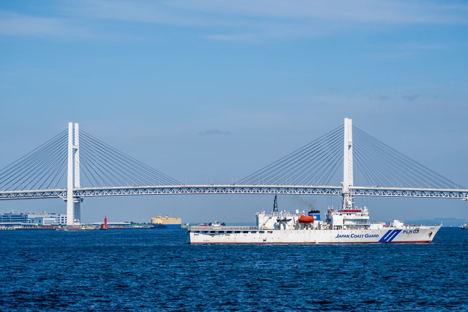 Japan Coast Guard vessel sailing in front of the iconic Yokohama Bay Bridge under a clear blue sky.