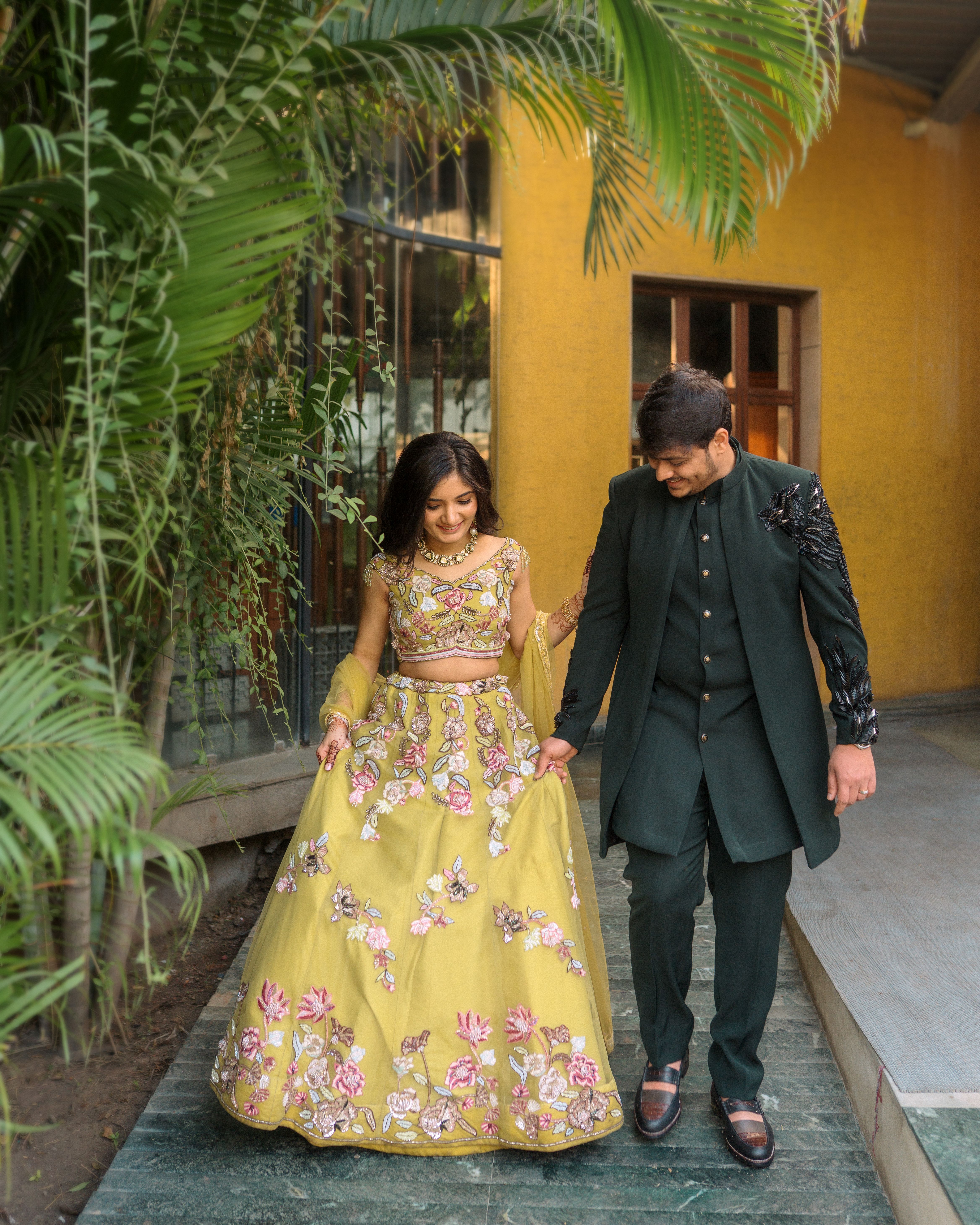 A happy engaged couple in vibrant Indian attire walking hand in hand outdoors.