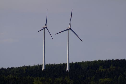 Two wind turbines stand tall against a backdrop of lush German forest under a clear sky.