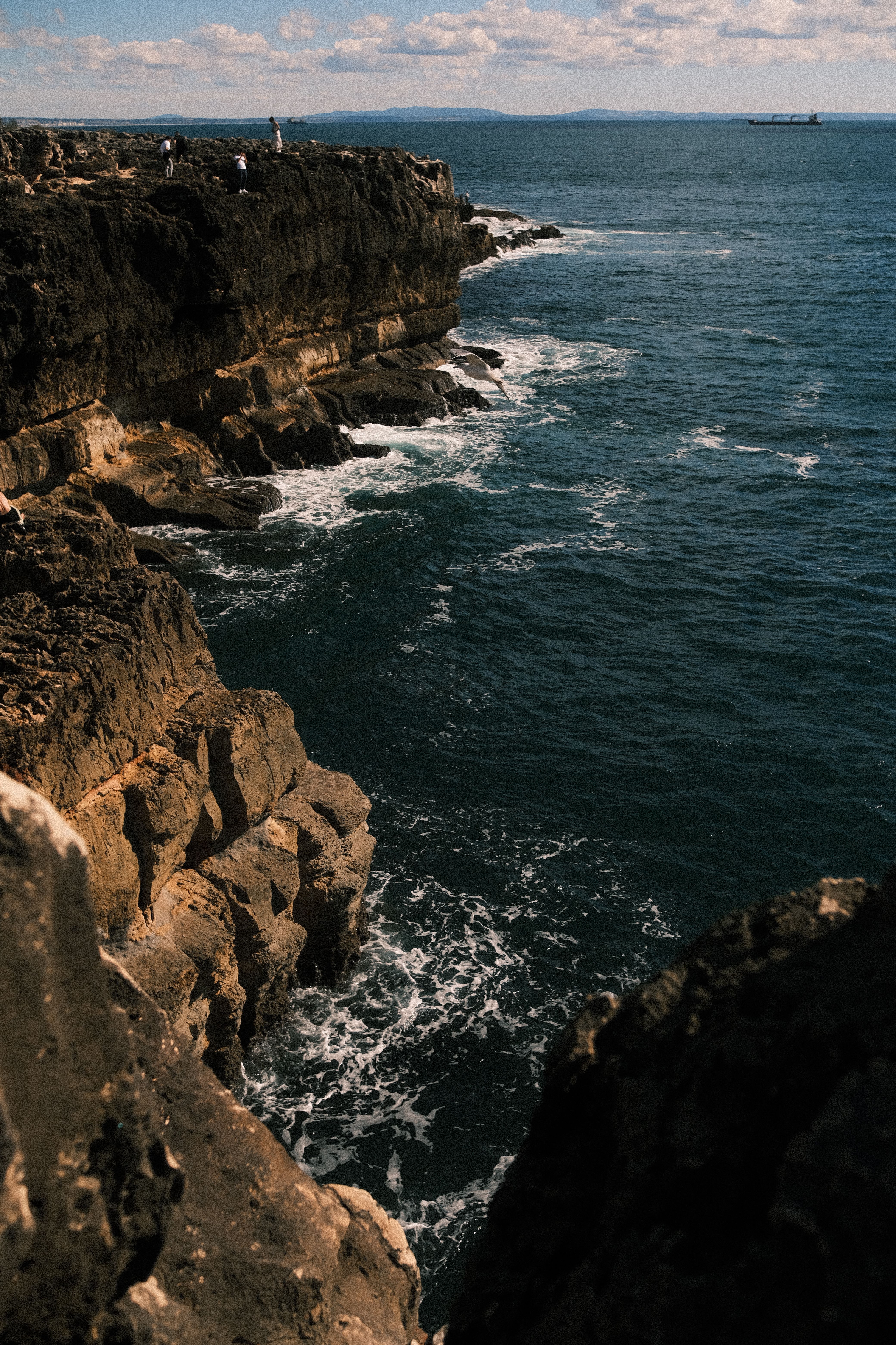 A dramatic seaside cliff with waves crashing along the rocky shore under a blue sky.