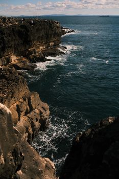 A dramatic seaside cliff with waves crashing along the rocky shore under a blue sky.
