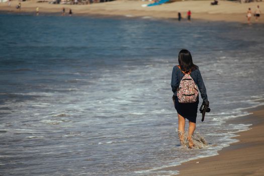 A woman with a backpack walking through gentle beach waves, capturing a relaxing seaside moment.