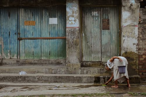 Free An elderly woman cleans the street outside aged blue doors in Kanchrapara, India, illustrating daily life. Stock Photo