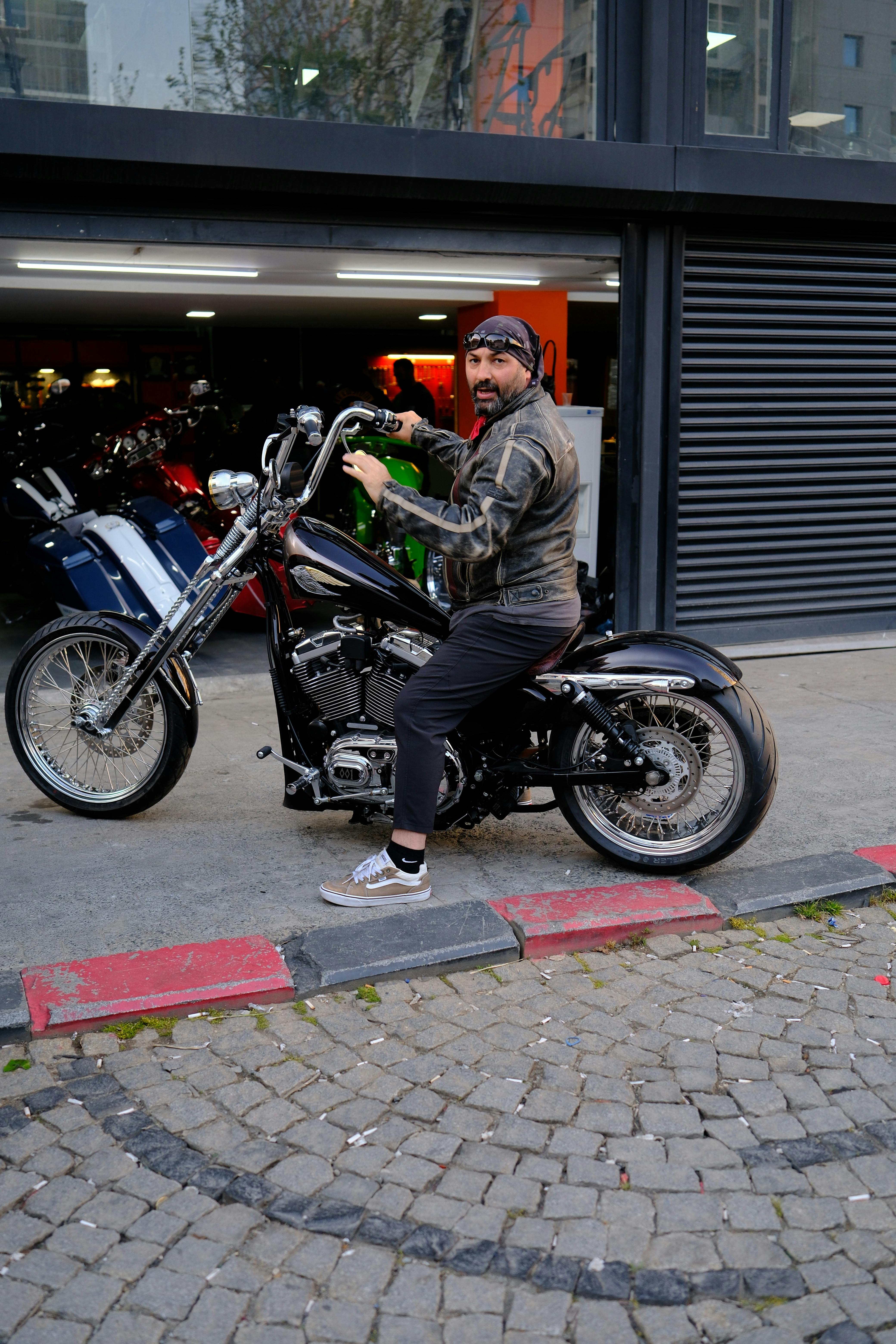 Man in leather jacket sits on chopper motorcycle outside a shop, showcasing biker culture.