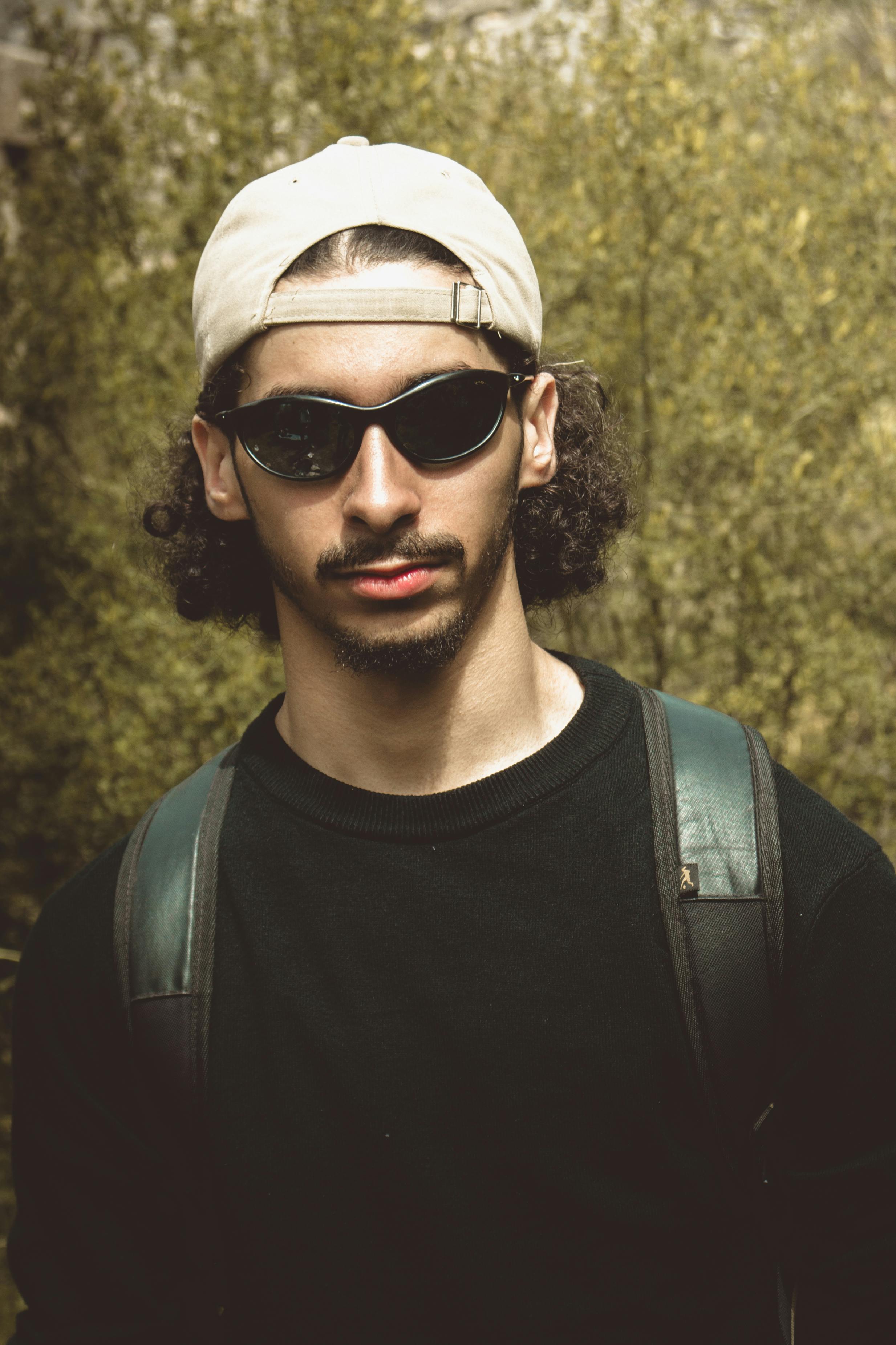 Stylish young man outdoors wearing sunglasses and a cap, carrying a backpack in El-Hamdania, Algeria.
