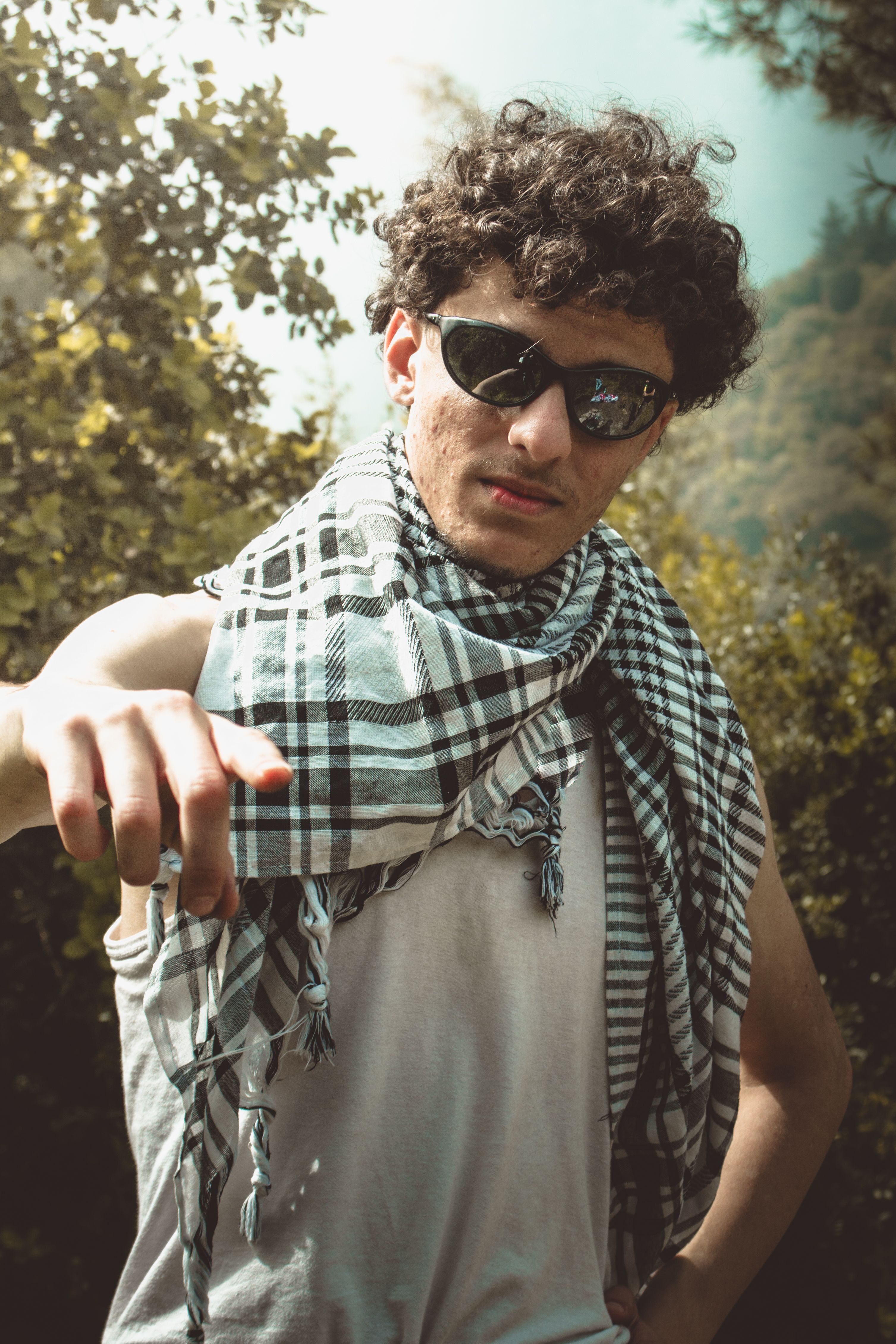 A young man wearing sunglasses and a plaid scarf poses confidently outdoors on a sunny day.