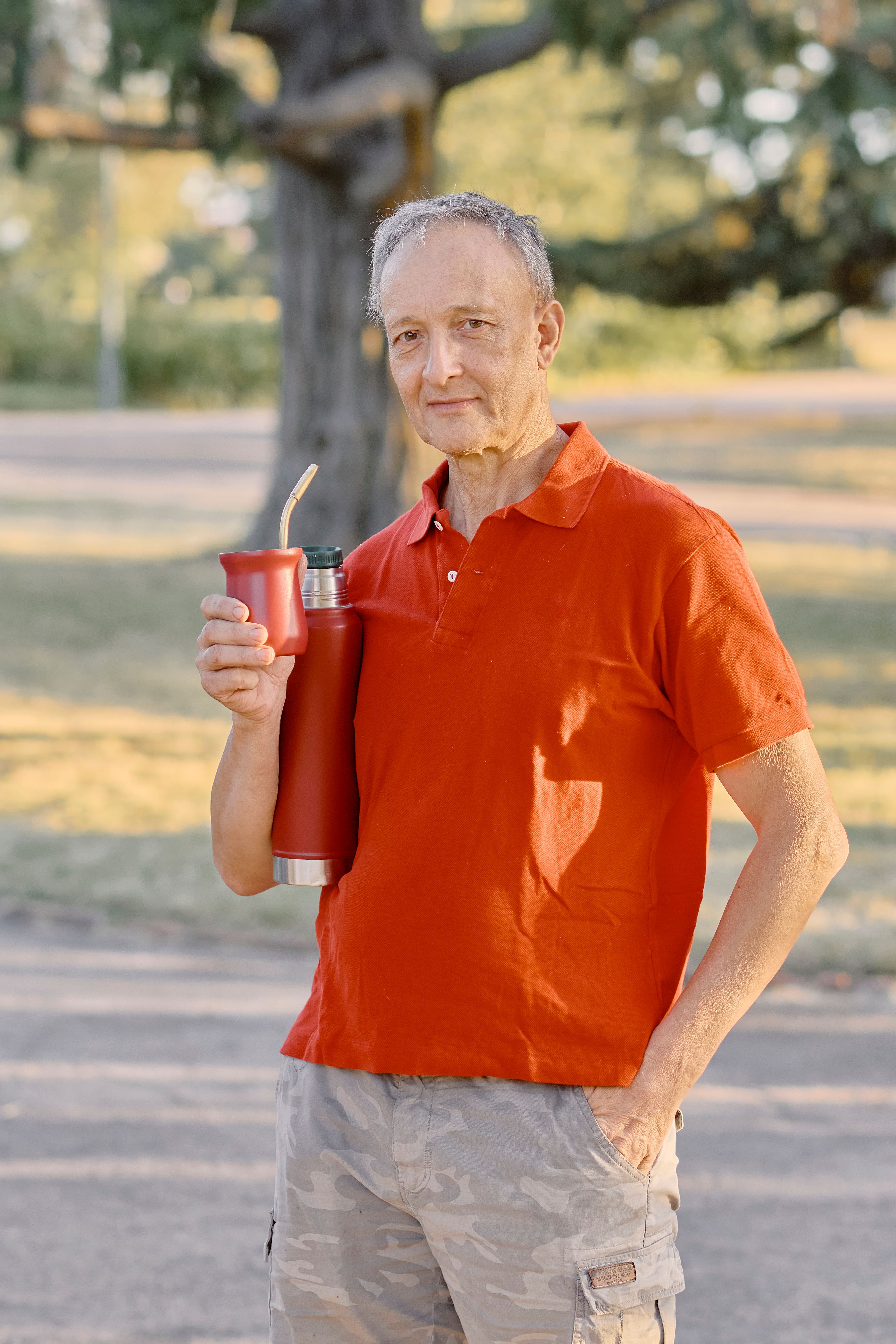 Senior man in red shirt enjoying yerba mate outdoors in a sunlit park, conveying a relaxed lifestyle mood.