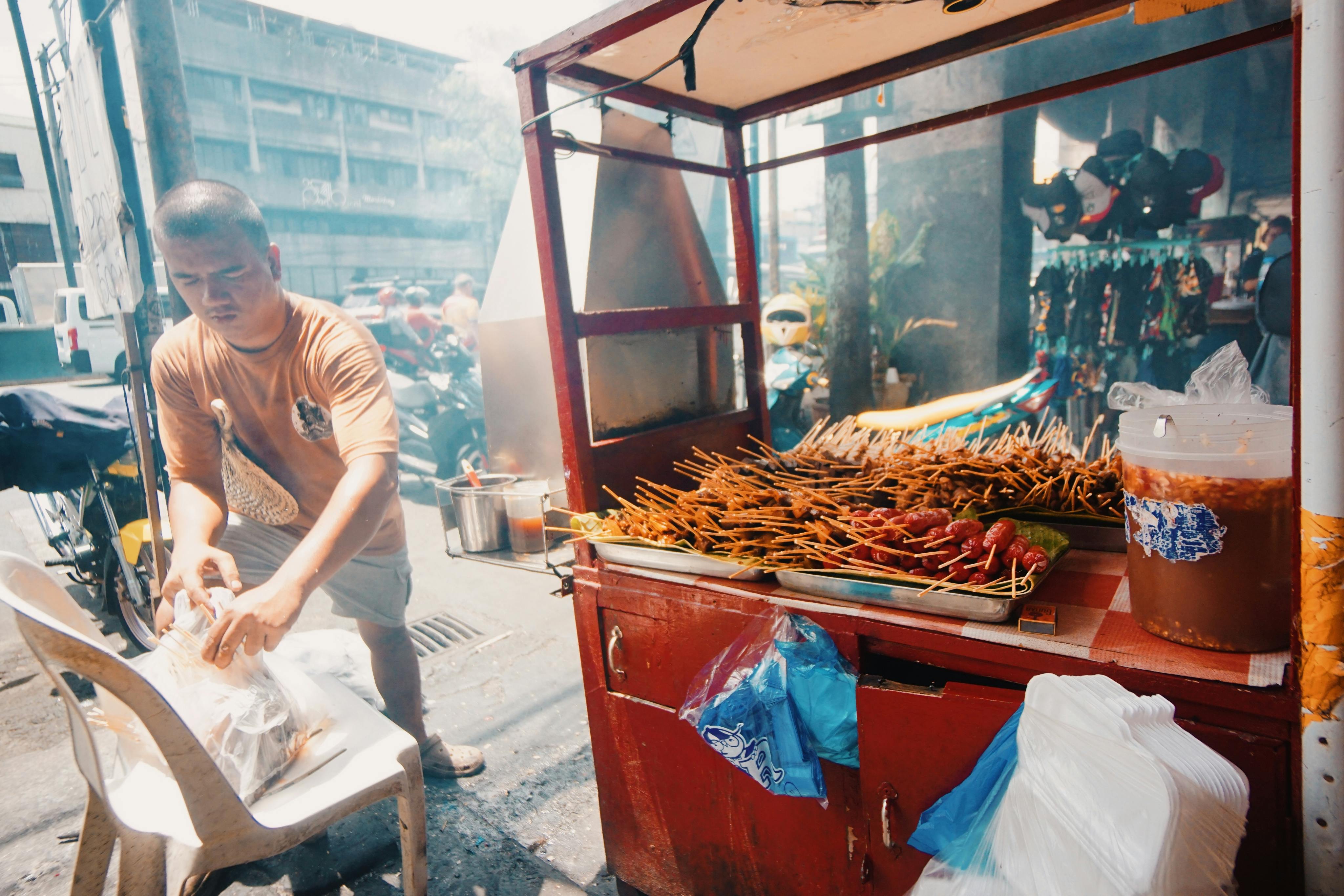 Asian street vendor prepares grilled skewers at a bustling outdoor market scene.
