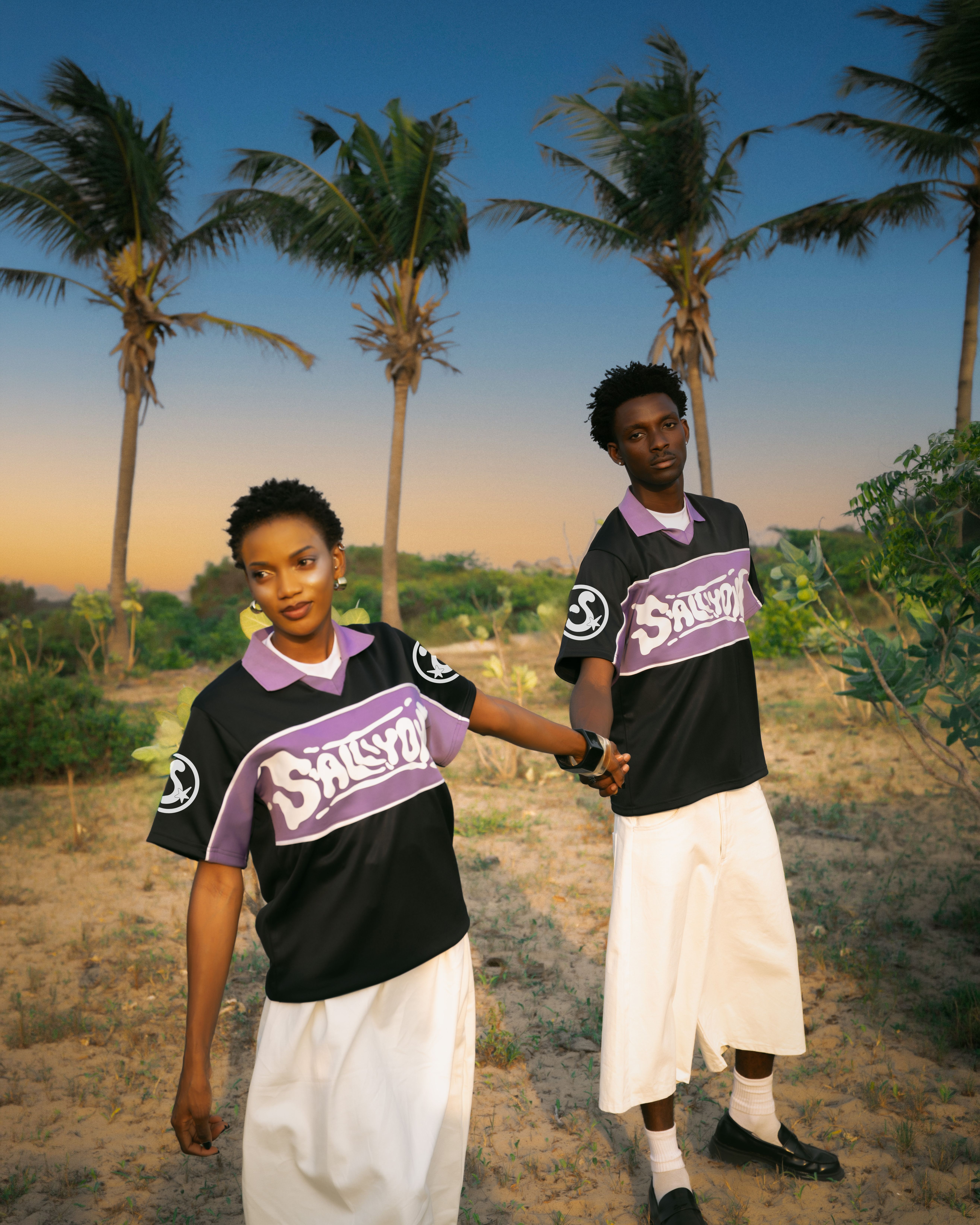 Fashionable couple in matching outfits pose among palm trees in a tropical setting.