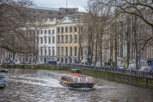 A scenic view of Amsterdam's historic canal with a tour boat surrounded by classic architecture in winter.