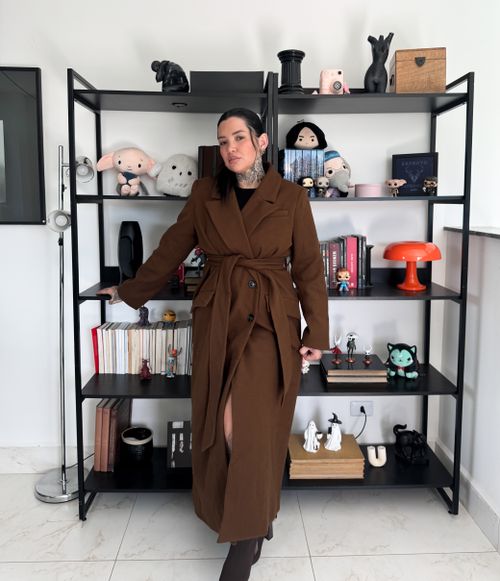 Free Fashionable woman posing in a brown trench coat in a modern home interior with bookshelves. Stock Photo
