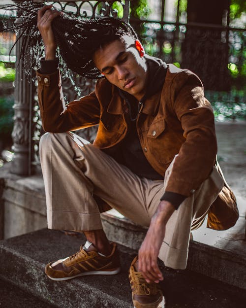 Stylish young African American man in sneakers and trendy jacket holding long braided hair with eyes closed