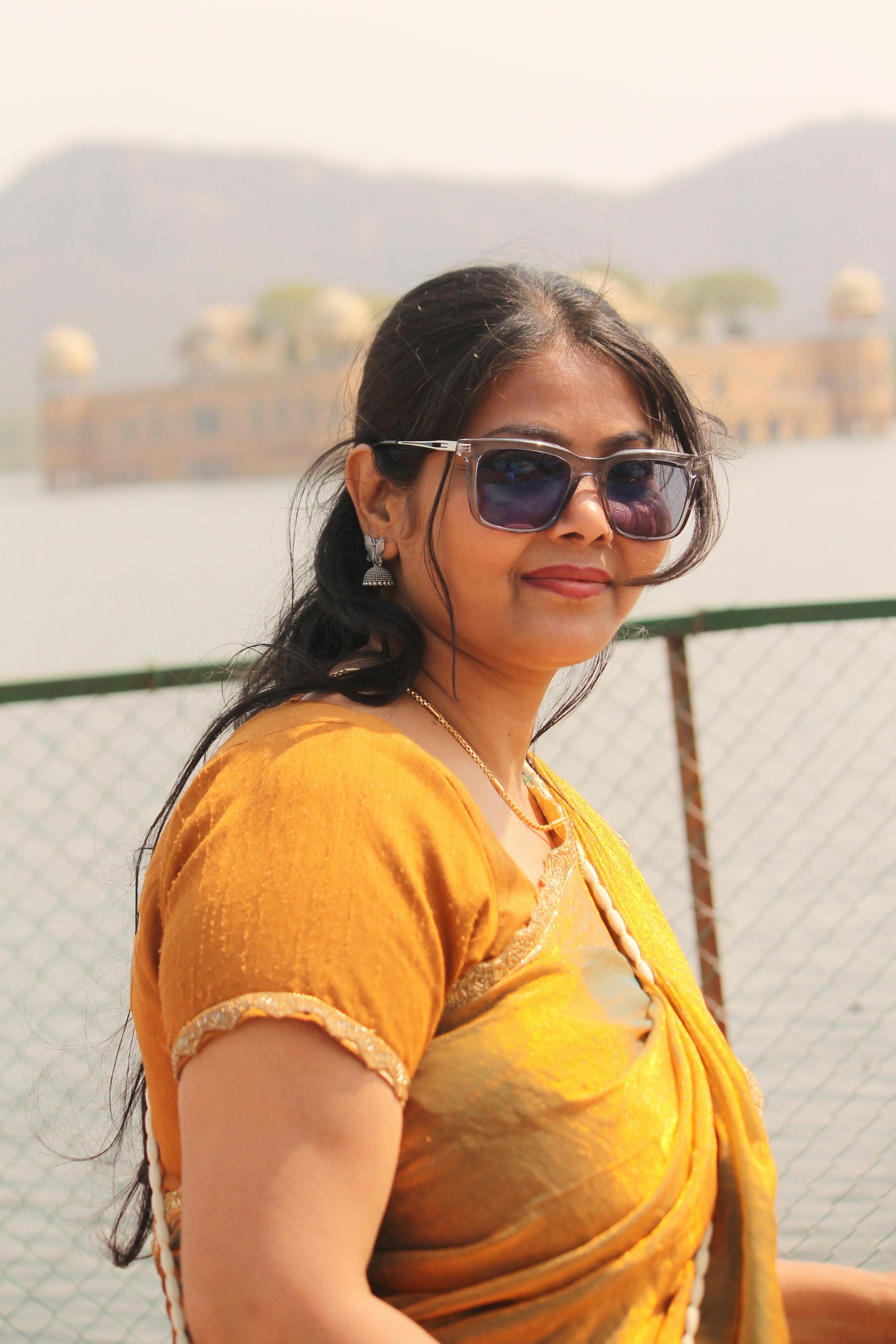 A woman in sunglasses and yellow sari smiles with Jal Mahal in the background.